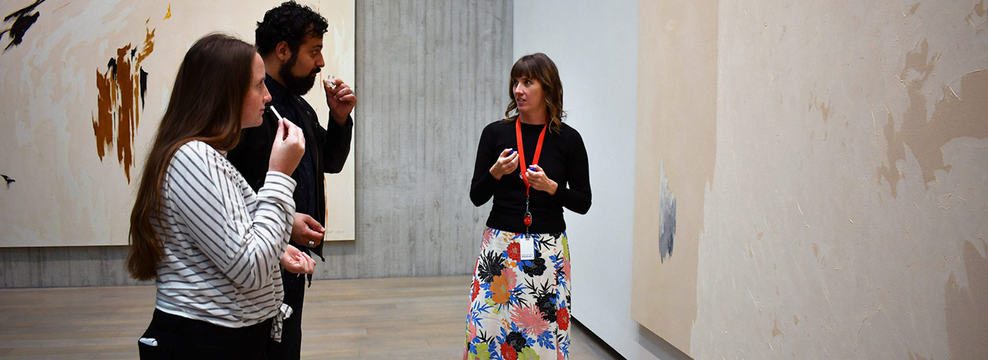 Two people smell items while a tour guide talks to them in a gallery