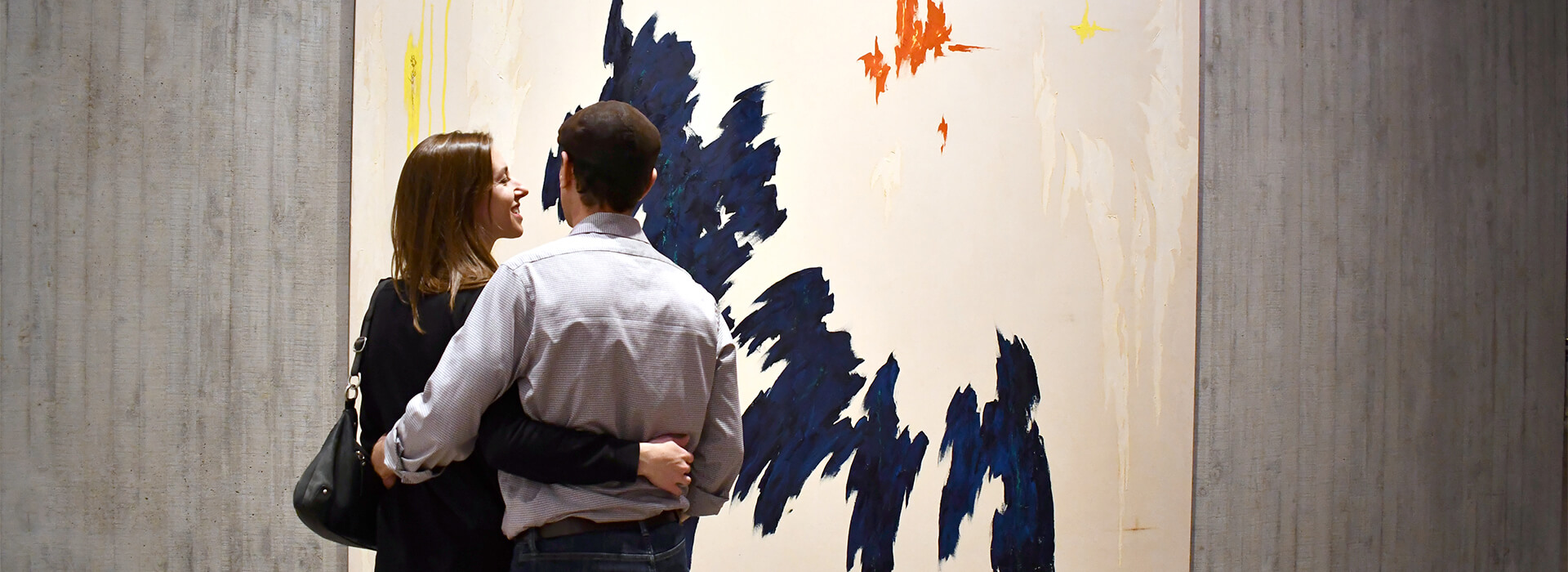 Woman smiles and looks at man as they put their arms around each other in front of a painting