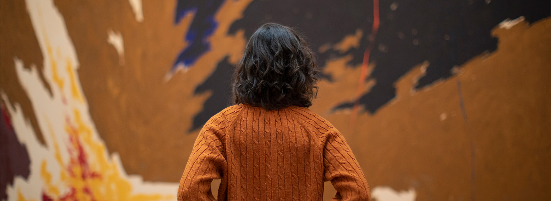 Woman wearing an orange sweater looks up at a large abstract painting with brown, yellow red, blue, black, and orange paint