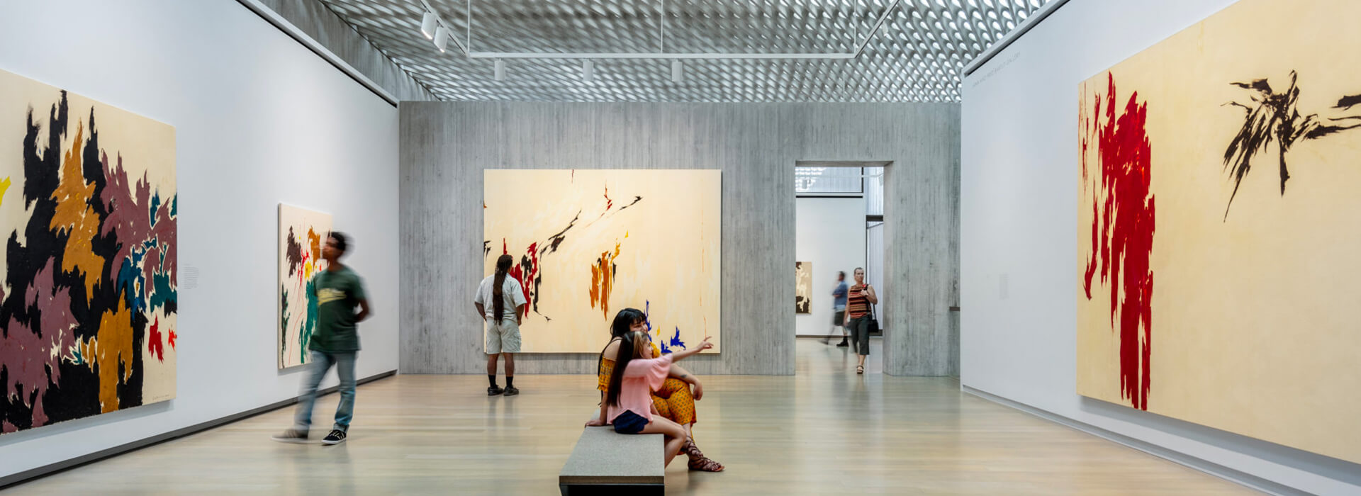 A mom and daughter sit on a bench while two men stand on the other side of the room looking at art
