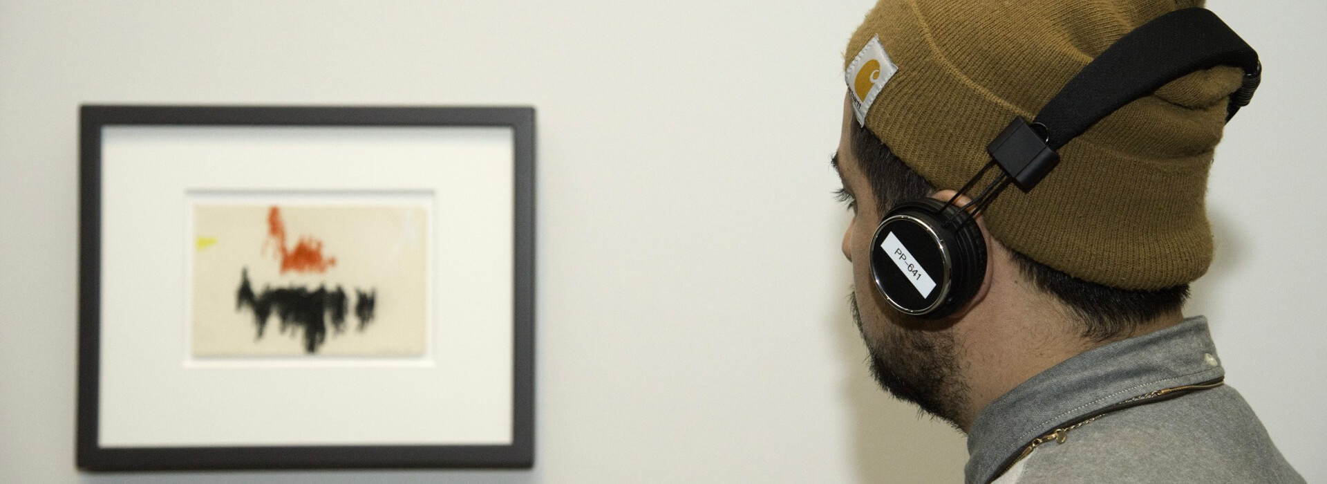 Man wearing headphones and a hat looks at a small framed artwork