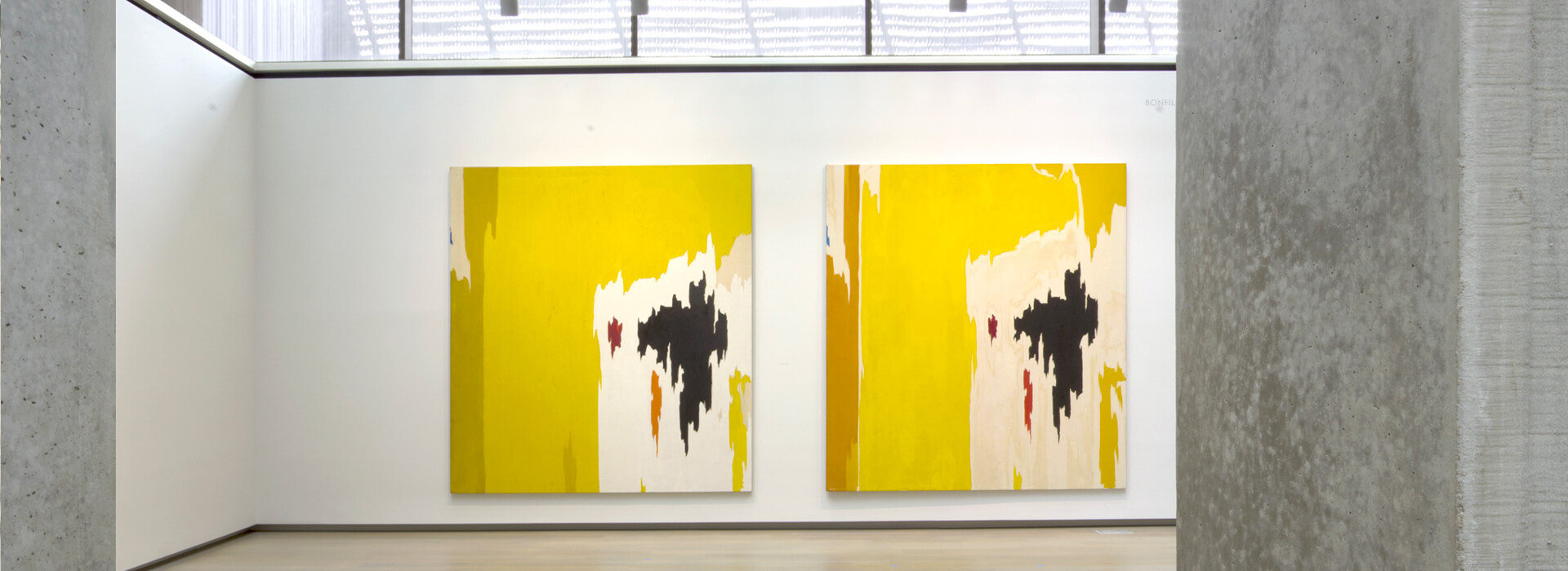 Two similar looking abstract oil paintings with mostly yellow, white and black paint hang on a wall next to each other