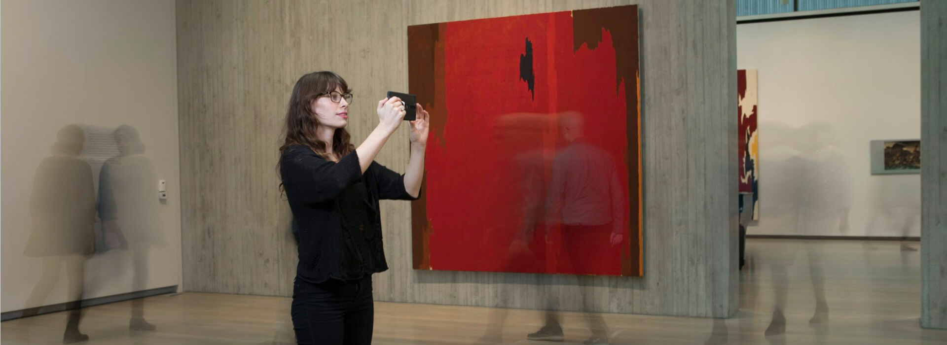 Woman holds up a cell phone and takes a photo of a painting with it while blurred figures appear around her