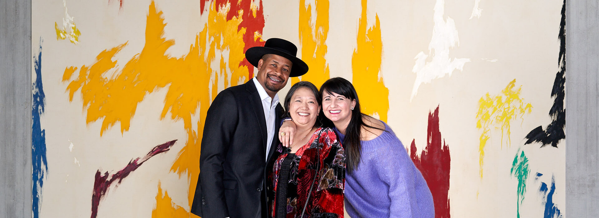 Two women and a man wearing a hat pose in front of a large colorful abstract painting