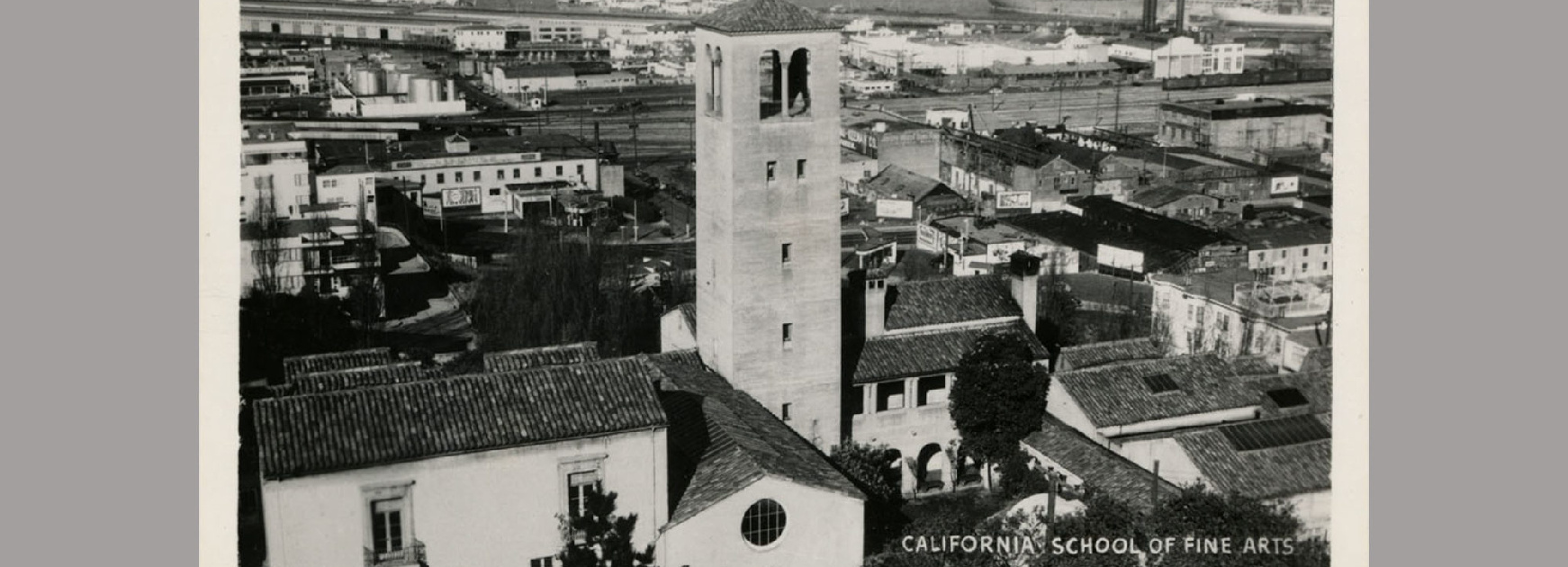 A black and white postcard showing an aeriel view of a group of buildings next to a body of water.