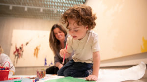 A young boy sits on the floor of a gallery and colors on a green piece of paper while his mom smiles behind him