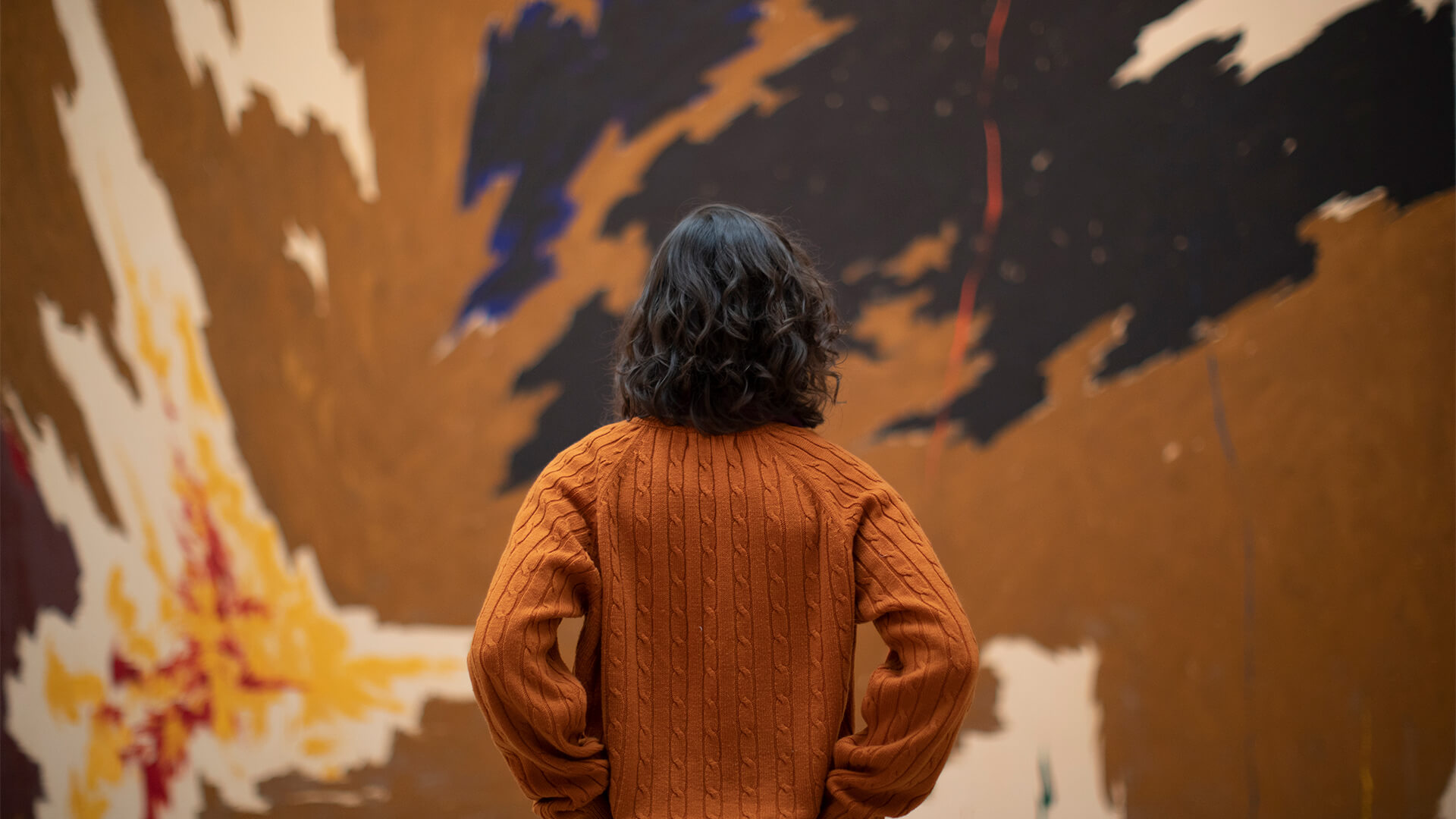 A woman in an orange sweater looks up a a large abstract painting by Clyfford Still