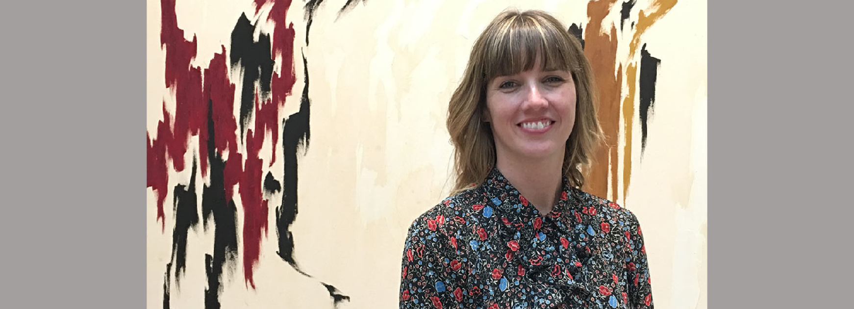 A woman with blond hair and bangs stands smiling in front of an abstract art piece.
