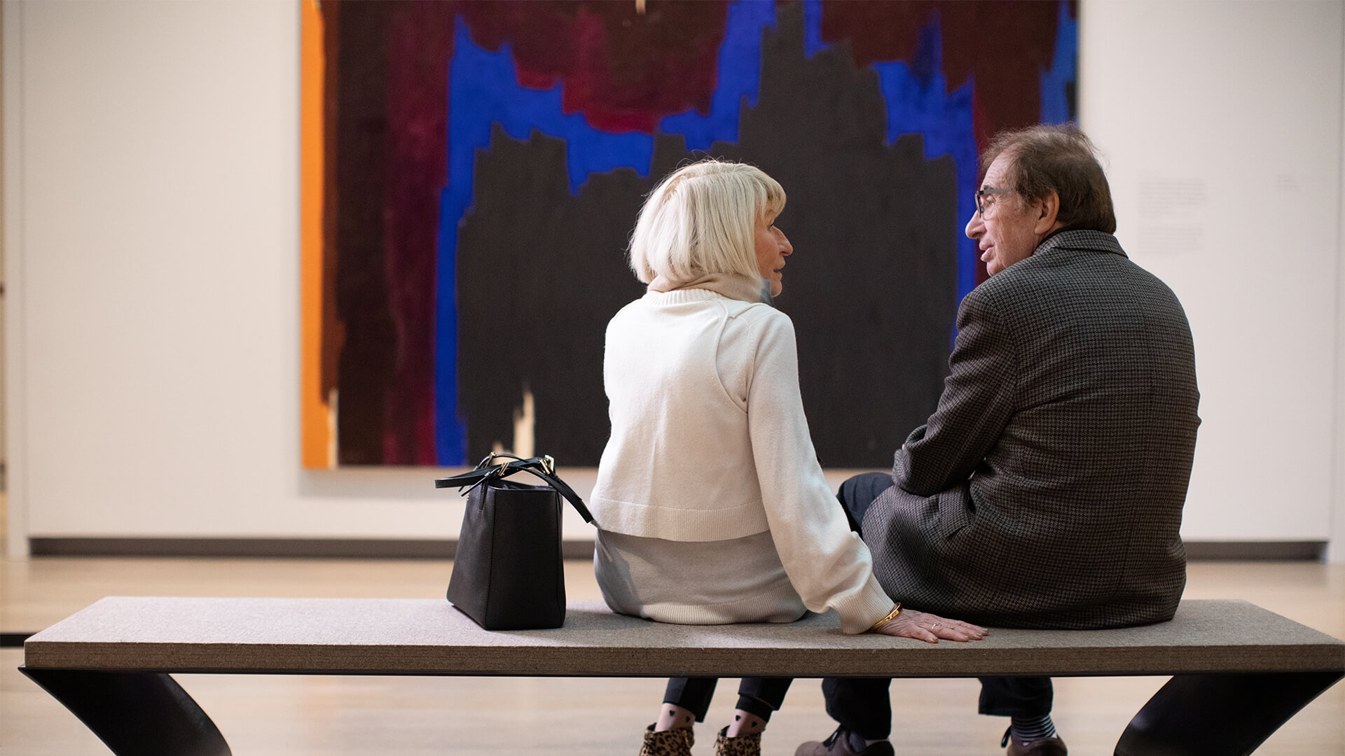 An older couple sits on a bench and looks at each other with a large colorful abstract painting in front of them
