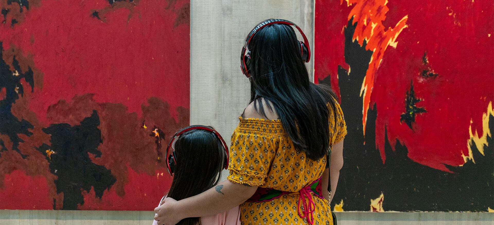 A mom puts her arm around her daughter's shoulder as they both wear headphones and look at two red and black abstract paintings on the wall