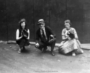 A black and white photograph of a man wearing glasses, a hat, and tie crouching down holding a baseball in one hand and a glove in the other and two women, one on either side, holding up baseball gloves