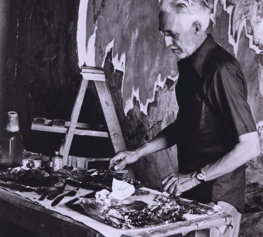 Clyfford Still looks down at a table with art supplies in his studio with a large oil painting behind him