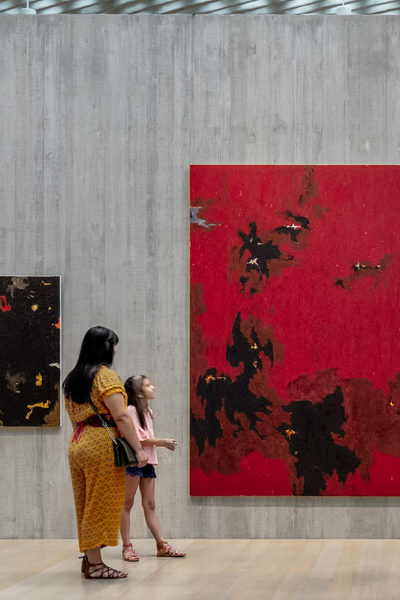 A woman and her daughter look up at a large red painting in a large gallery space