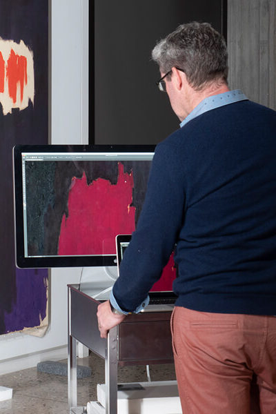 A man takes photos of a large abstract oil painting hanging on the wall with a monitor in front of him