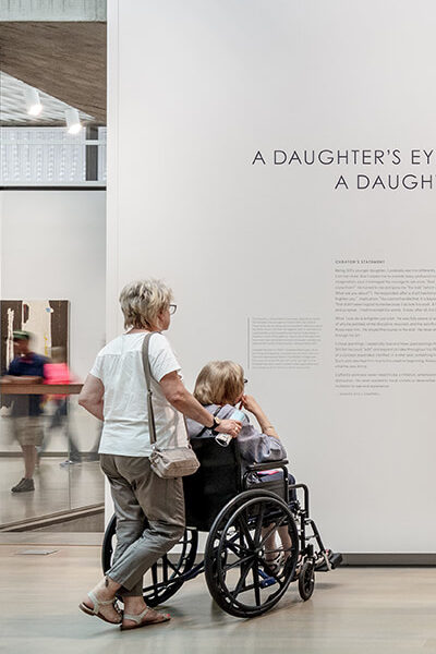 A man stands and looks at text on a wall next to a painting with a woman in a wheelchair and another woman pushing the wheelchair next to him