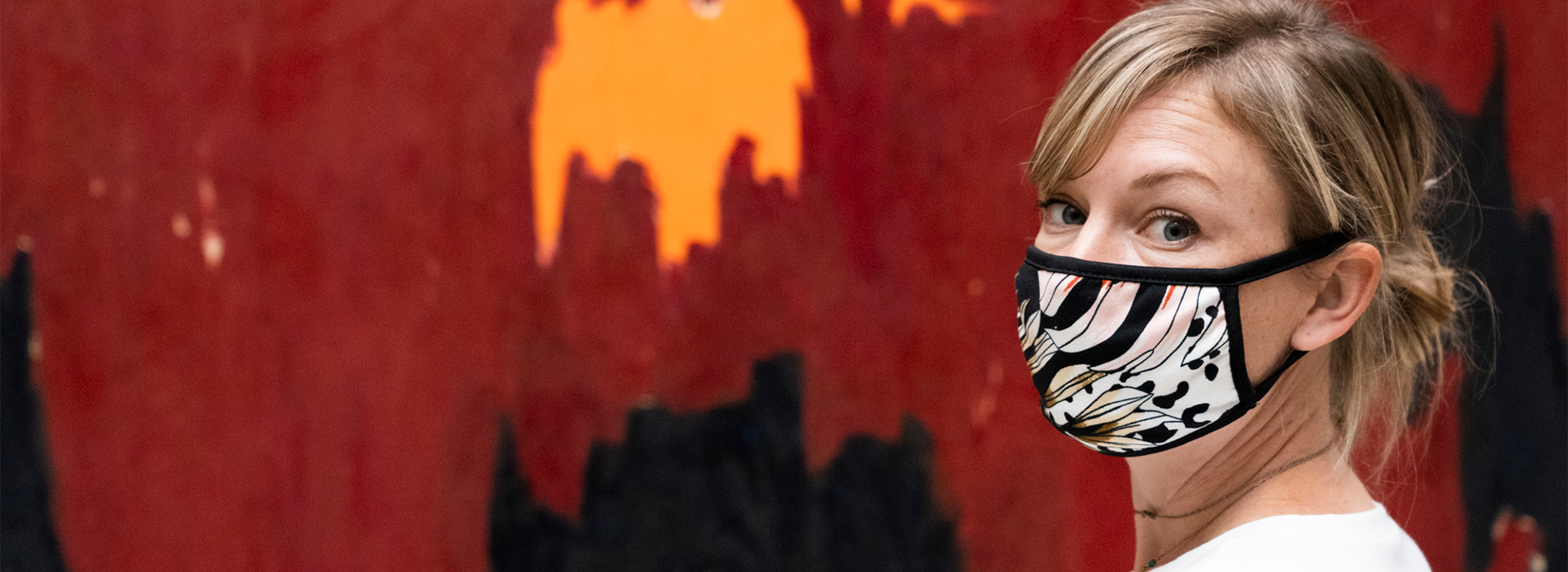 Woman wearing a face mask with a pattern looks at the camera with a red Clyfford Still painting in the background