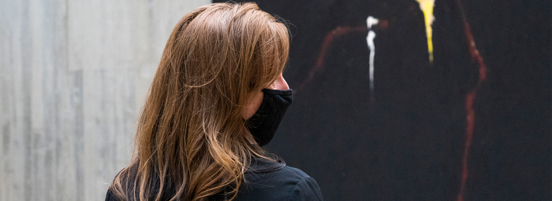 Side profile photo of a woman wearing a mask standing in front of a mostly black abstract painting by Clyfford Still
