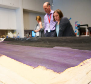 Two women and a man look down at an unrolled painting on canvas with purple and black paint