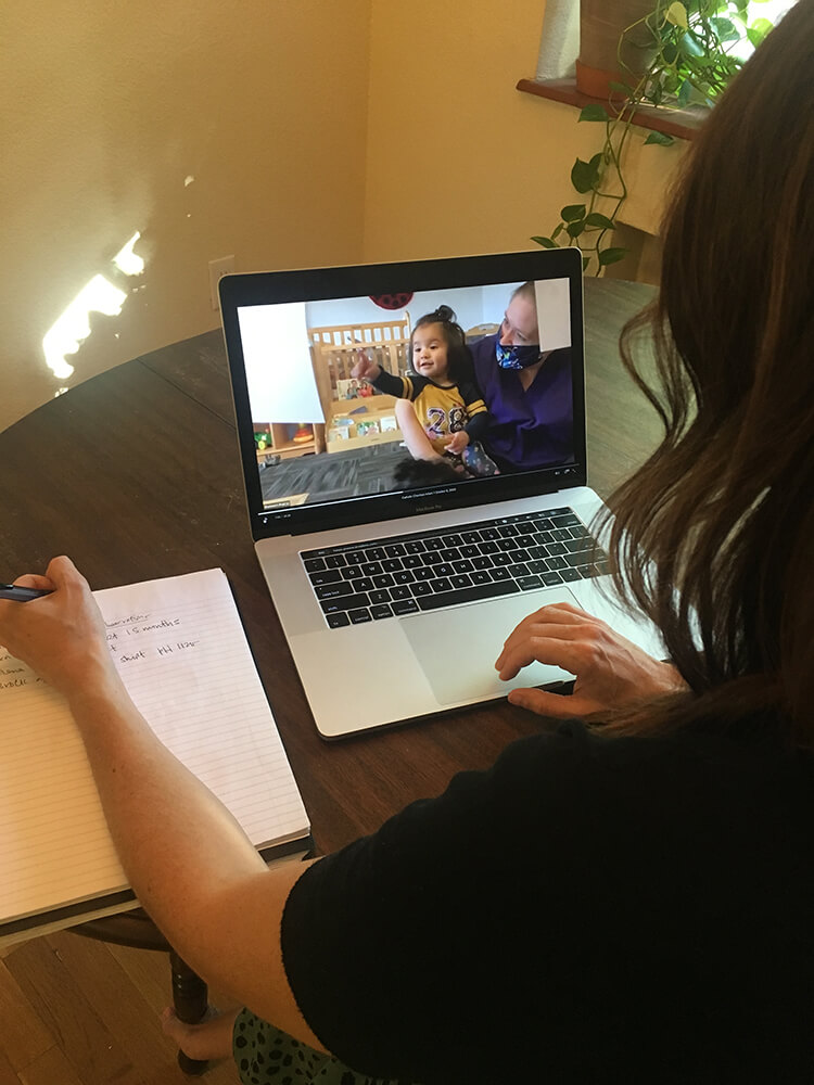 Woman looks at a laptop with children and a teacher on the screen and she writes something in a notebook