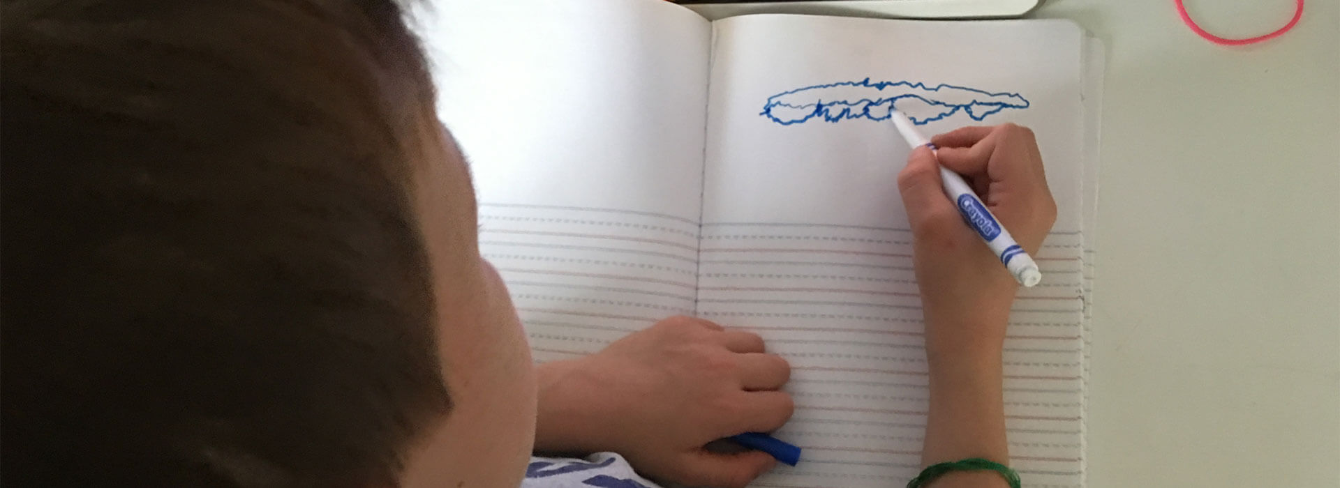 Looking down as a student draws an abstract shape in a notebook using a blue marker while looking at a tablet