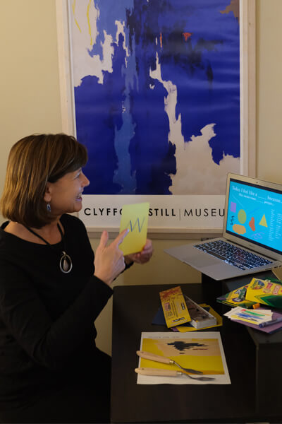 A woman holds up a card directed at a laptop with a blue Clyfford Still painting poster on the wall behind her
