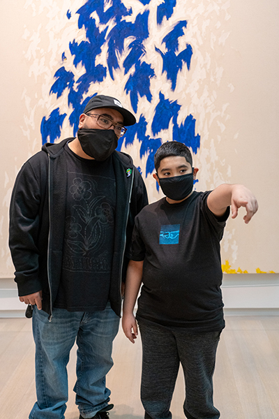 A father and son wearing masks look at art while the son points at something in the painting