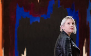 Woman wearing a black leather jacket looks up into the distance as she sits on a bench in front of a colorful abstract oil painting with blue, black, orange, maroon, and white paint