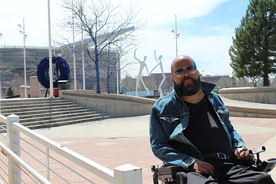 Man in a motorized wheelchair with a beard wears sunglasses and looks at the camera in front of large abstract sculptures of people