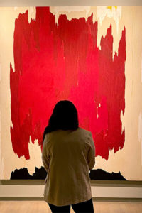 Woman has her back to the camera and looks up at a large abstract red painting