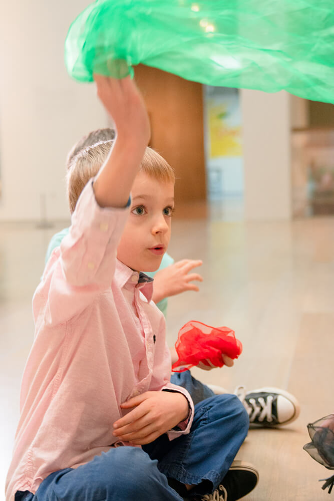 A young boy in a pink shirt waves a green scarf in the air while sitting on the floor of a museum gallery