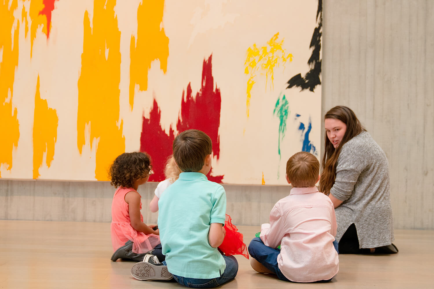 A group of young children sit on the floor in front of a large colorful painting with a teacher