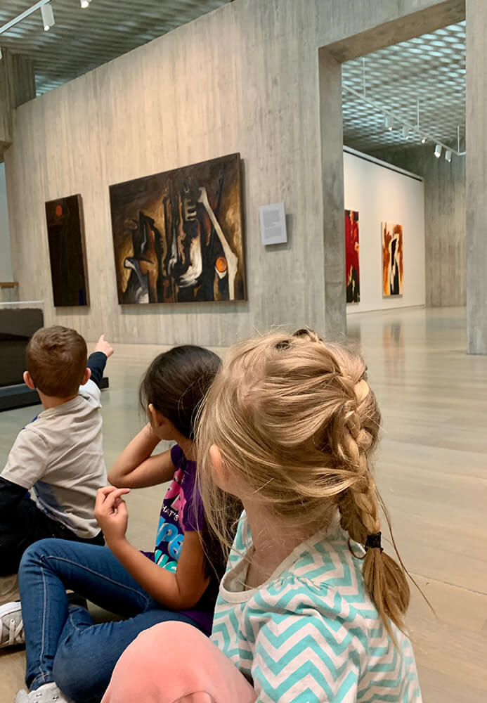 Young children sit on the floor and turn their heads to look at art on the wall of a museum gallery