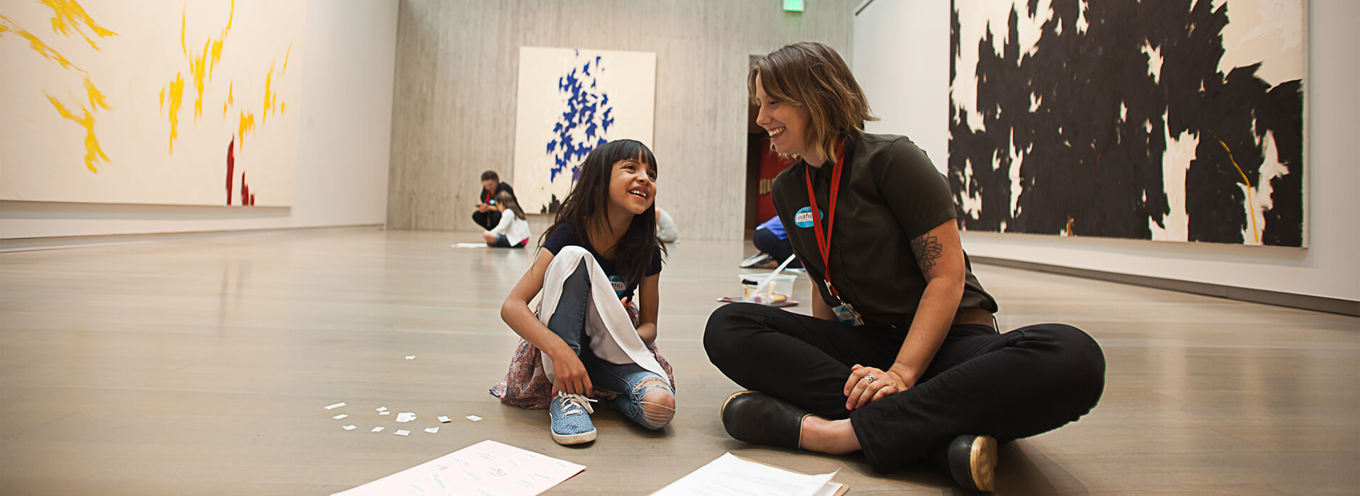 A young student sits on the floor of a museum gallery and smiles up at a teacher, also seated on the floor