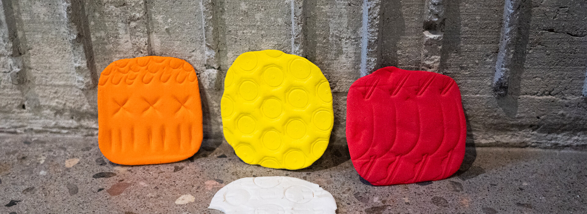 Orange, yellow, red, and white clay patterns lean up against textured concrete