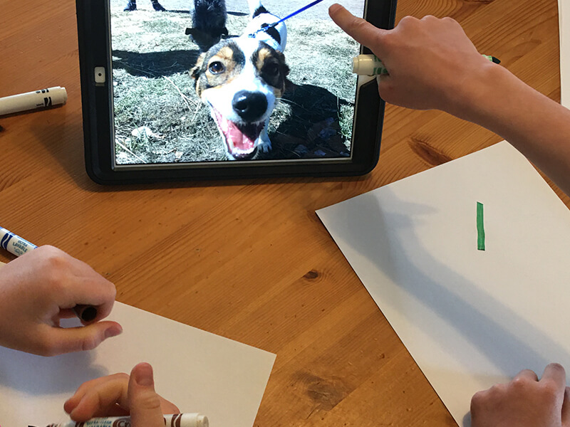 Two children with paper and markers look at a photo of a dog and draw the outlines