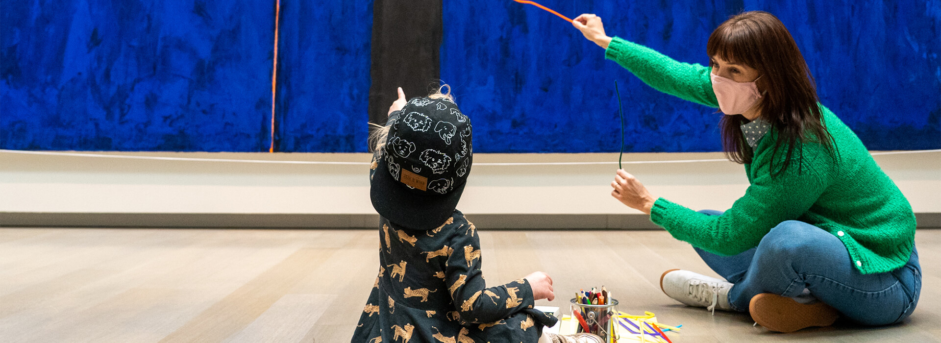 A woman wearing a green sweater and a mask sits on the floor and points up at an abstract blue painting with a pipe cleaner and a young child sits next to her and points up with her finger at the same spot