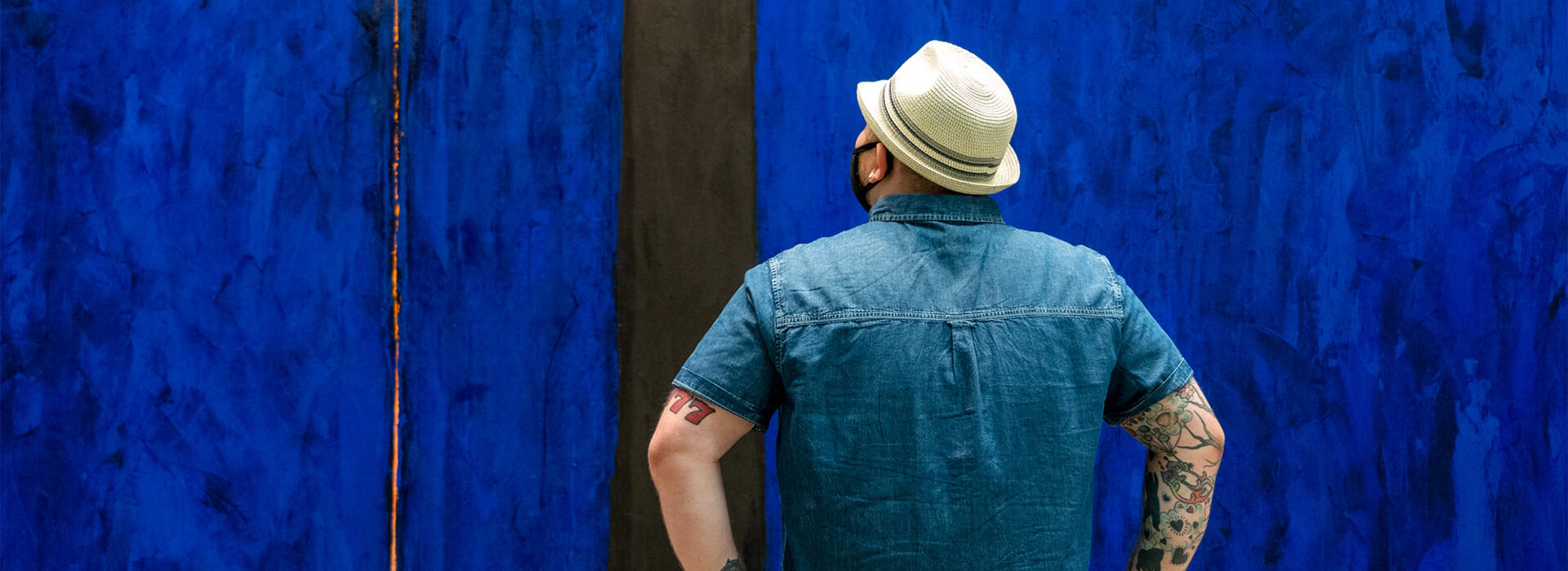 Man wearing a blue short sleeve shirt and a white hat looks up at a large blue abstract painting