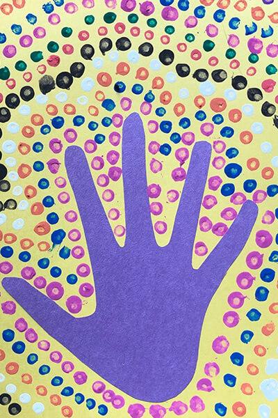 Purple hand made out of construction paper with painted dots all around it on a yellow piece of paper