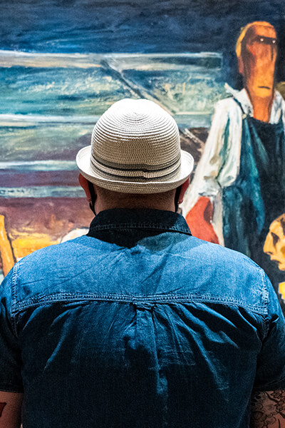 Man wearing a blue shirt and a white hat looks up at an abstract painting of farmers