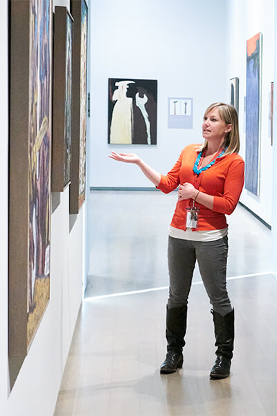 A woman wearing an orange sweater gestures at paintings on a wall to a tour group