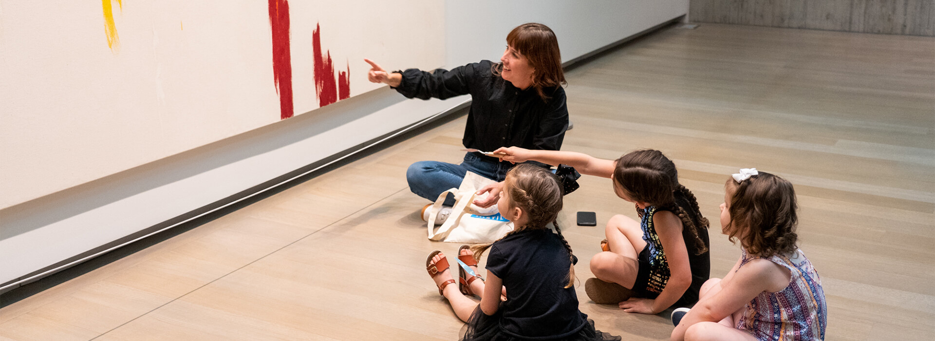 A teacher sits with three young girls on the floor in front of a painting and they point at a spot on the painting