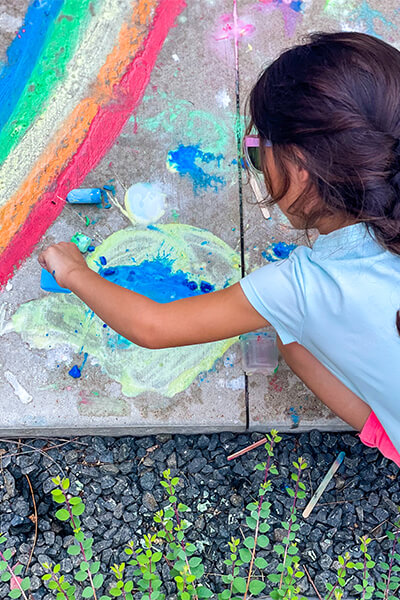 Girl paints on the sidewalk with colorful ice chalk and makes a rainbow