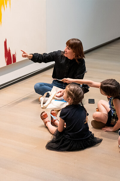 A teacher sits with two young girls on the floor in front of a painting and they point at a spot on the painting