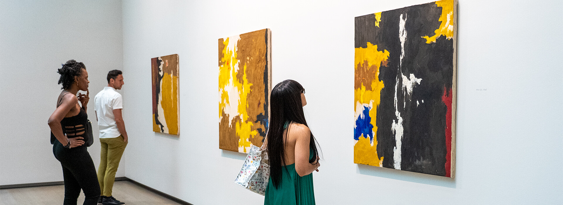 A woman wearing a green dress, another woman dressed in black top and black pants, and a man wearing tan pants and a white shirt all look pensively at three abstract paintings on a wall