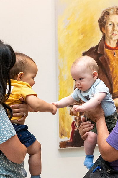 Two babies being held by women reach for each other in front of a painting