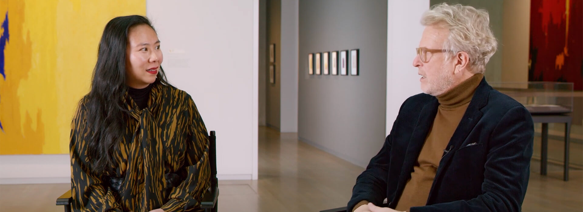 Two people sit in chairs in an art gallery and talk to each other