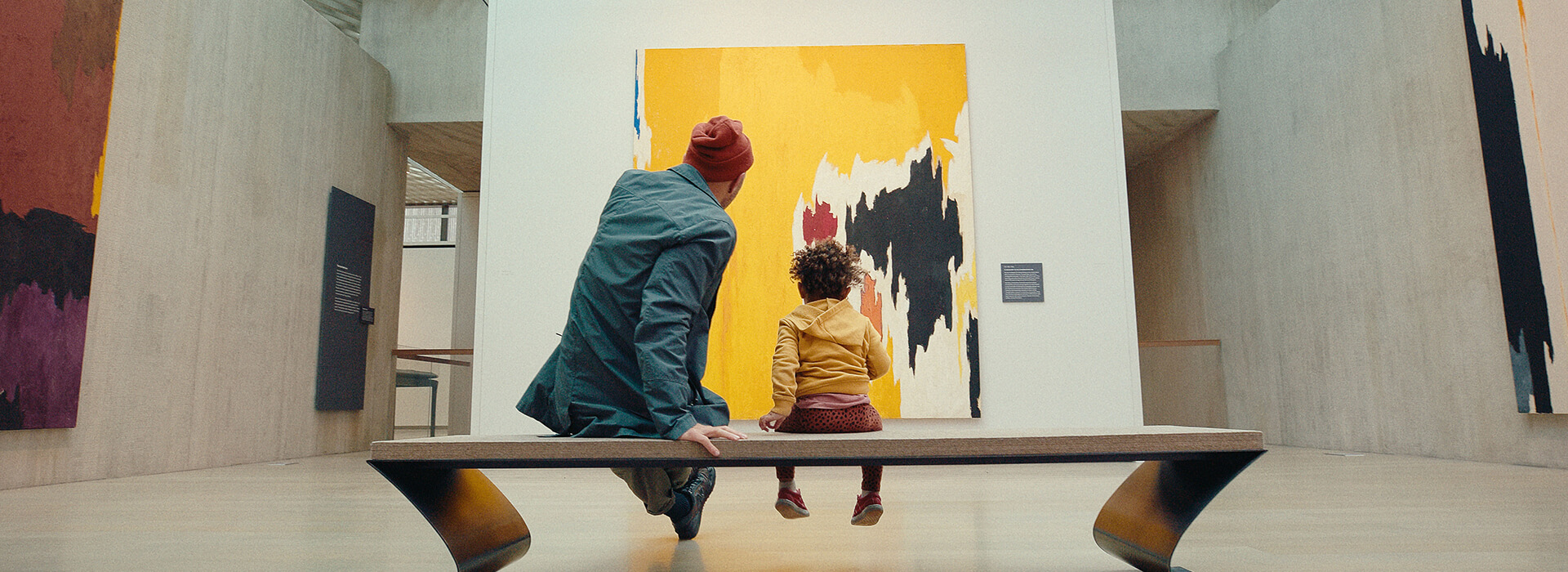 A father sits next to his toddler son on a bench as they look at a large yellow abstract painting