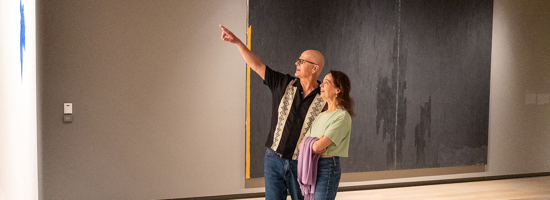 A man points up at a painting while the woman next to him looks at it and smiles