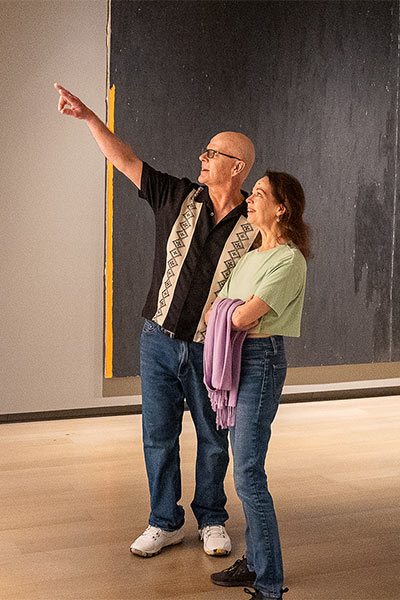 A man points up at a painting while the woman next to him looks at it and smiles