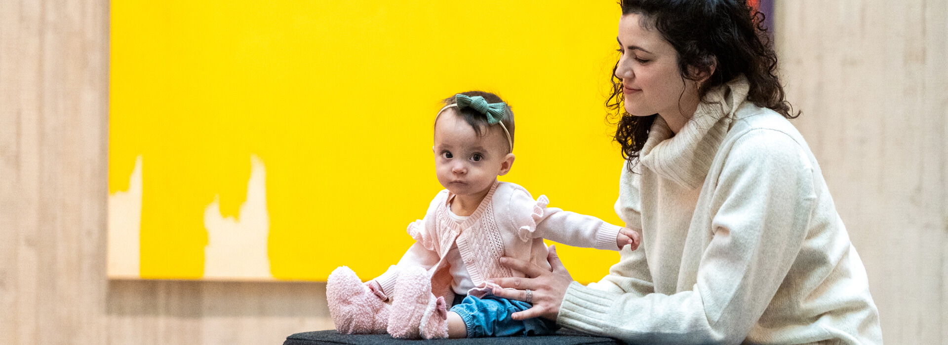 A mom kneels next to her baby girl who is sitting on a cushion in front of an abstract yellow painting